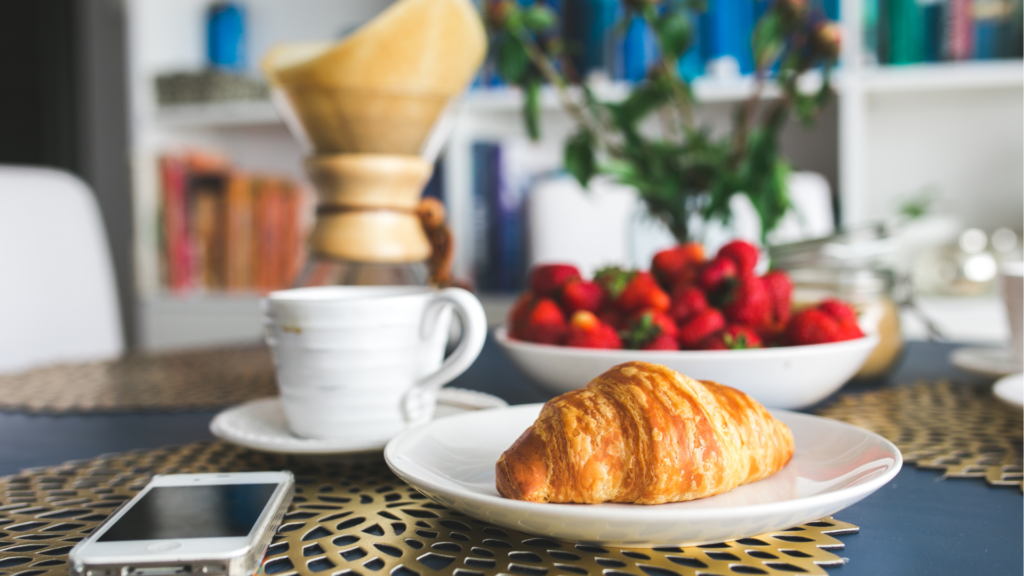 A peaceful breakfast scene with a fresh croissant, a cup of coffee, and a bowl of strawberries on a sunlit table, while a smartphone rests untouched nearby. The calm, intentional moment reflects a digital detox approach — choosing slow, mindful living over early-morning screen time.